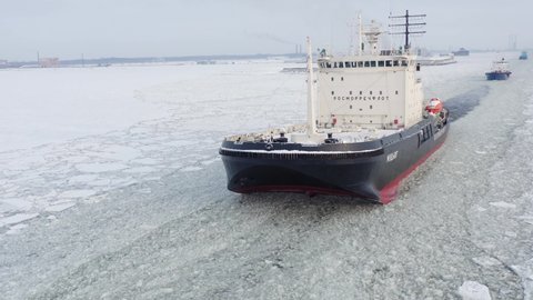 St. Petersburg, Russia, winter 2022: Icebreaker boat Mudug clear the way for carho ship. Frozen landscape around. Aerial view.