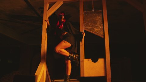 Young sexy woman in mask and black Halloween executioner costume dancing near old vintage wooden guillotine .  Girl with beautiful body with red hair wearing shorts dancing inside dark building . 