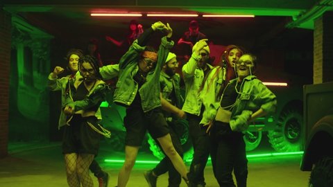 Group of stylish young people dancing , moving near military tank inside garage building Colorful stylish clothes , hippie style fashion . Modern dances . Purple led light , slow motion