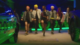 Group of stylish young people dancing synchronized dance  , moving near military tank and police car inside garage building Colorful stylish clothes  Modern professional dances . Green blue led lights