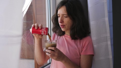 WROCLAW, POLAND - FEB 16, 2022: Woman pours a coca cola foaming sparkling  beverage in a glass and drinks it standing by a window at home