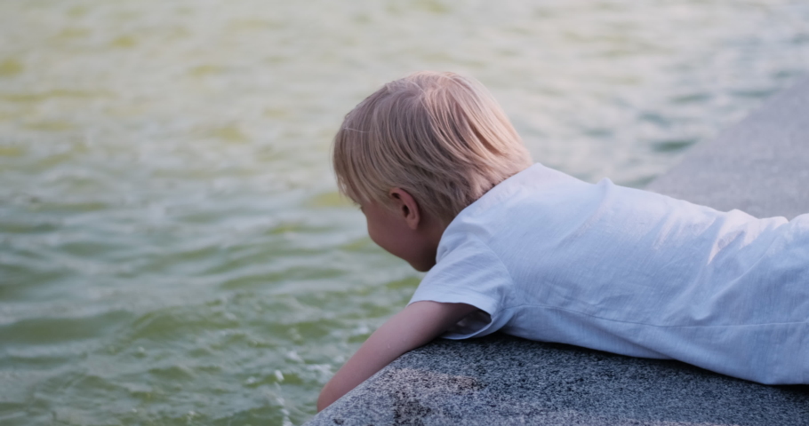 Boy lies on his stomach with hands dipped in the water. Child plays and makes water splashed.