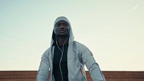 Athletic black man is stretching outdoor on a sunny day. He is wearing unbranded sports clothes and headphones. Brick wall and mountain landscape on a backgroung. 4k slow motion video.