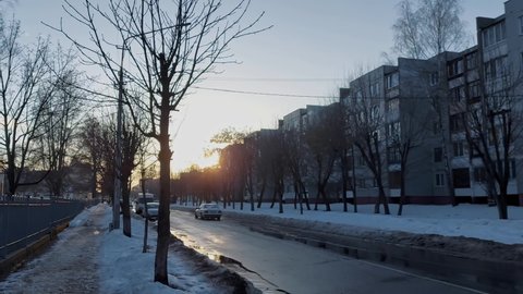 Car drives along a narrow street in winter in a residential area with multi-storey buildings in the evening during sunset. Early spring after winter. Above-zero temperature. Slush.