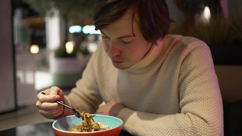 Young man in sweater enjoys eating food from ceramic bowl at table on blurred background. Guy has dinner in small cafe of supermarket close view