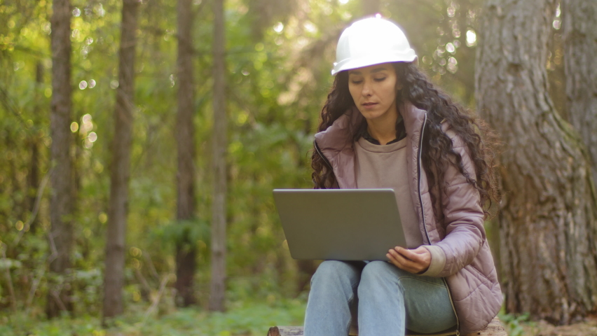 Millennial woman young experienced specialist forestry engineer environmentalist technician in hardhat checking trees entering data into laptop taking reforestation measures observing nature reserve