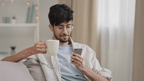 Young relaxed guy indian arabic bearded handsome man sitting at home on couch drinking tea coffee from cup looking at phone smiling watching funny video online chatting browsing with smartphone