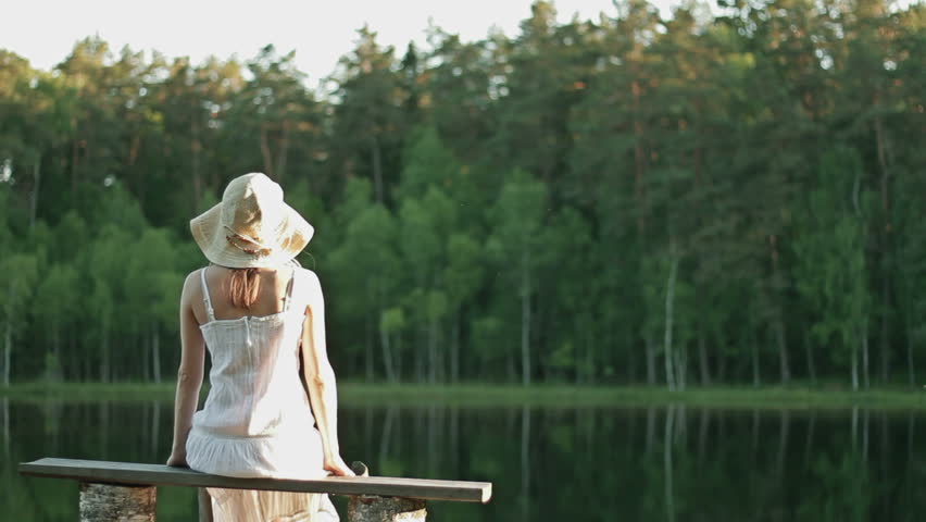 Happy romantic woman sitting by lake, wearing white dress, rear view
