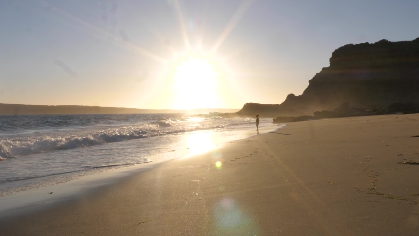 Beautiful shot of a young boy jumping in waves on a beach with the sun setting behind him.