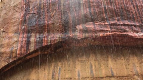 Rain pours down in front of a red sandstone arch in Zion national park that is streaked with black, red, copper and yellow color where water oxidizes the minerals in the stone. No audio