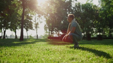 Attractive stylish man sitting in squat in park. Relaxed guy look in distance. Joyful person lean on knees resting in summer sunlight. Men inspecting city garden alone. Outdoor daily walk in garden