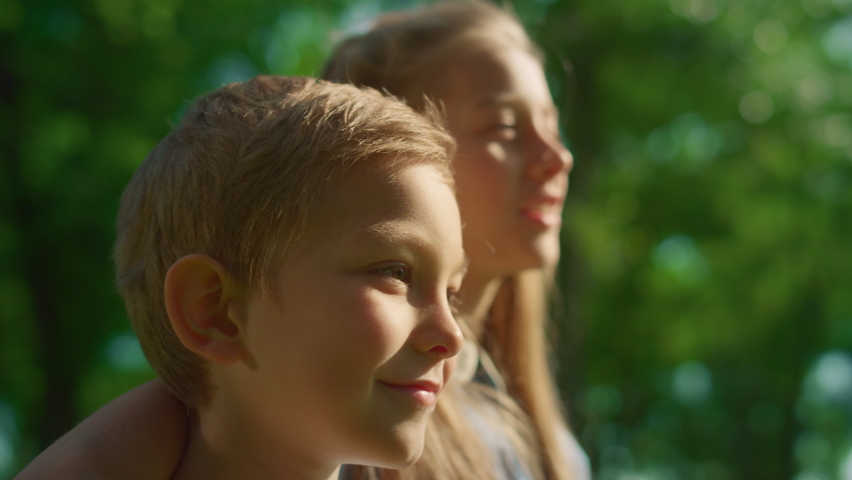 Two smiling kids stand lookin in distance. Blond siblings hugging close up. Joyful brother sister chatting in golden sunlight. Children pose in summer park on family picnic. Carefree childhood concept