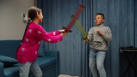 Multiethnic diverse kids playing active fight games at home. Cute black boy, charming girl fighting with toy swords in domestic room. Preteen contemporary friends having fun indoors. Leisure activity