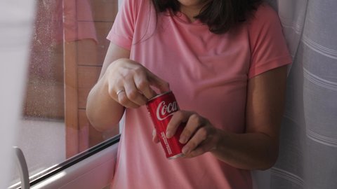WROCLAW, POLAND - FEB 16, 2022: Brunette woman portrait opens up a coca cola can and drinks carbonated beverage while standing by the window at home