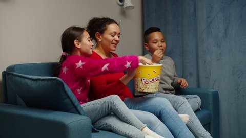 Single mother bringing popcorn bucket sitting down to couch, watching movie, TV show, online service with daughter and son indoors. Multiethnic family with two children having pastime together at home