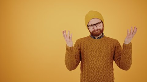 middle-aged bearded millennial man in a beanie and warm yellow sweater throwing hands in the air not knowing what to do medium studio shot yellow background. High quality 4k footage