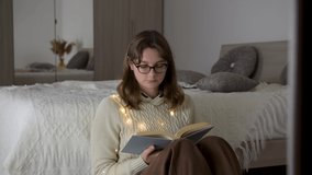 4k Young woman in eyeglasses, wearing sweater, is reading book in cozy hugge room with pampas grass, turning page, learning, studying, education, relaxation. Cozy winter day at home.