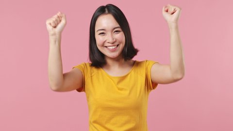 Excited jubilant overjoyed young woman of Asian ethnicity 20s wears yellow t-shirt doing winner gesture celebrate clenching fists say yes isolated on plain pastel light pink background studio portrait