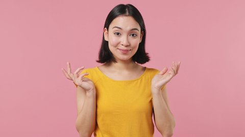 Confused shy shamed young woman of Asian ethnicity 20s wear yellow t-shirt look camera spreading hands say oops ouch oh omg i am so sorry isolated on plain pastel light pink background studio portrait