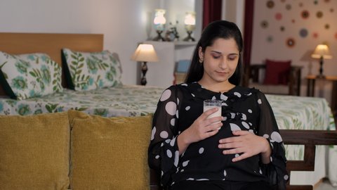 A pretty pregnant woman drinking a glass of milk - nutritious food. An Indian expectant lady gently stroking her hand on her stomach while sitting on a comfortable sofa - future parent, baby bump