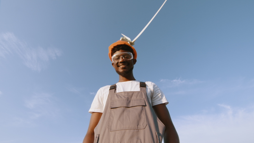 African american technician in helmet, glases and overalls looking at camera while standing on field with wind turbines. Concept: renewable energy, technology, electricity, service, green, future