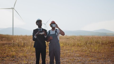 African american businessman and indian engineer wearing VR headset during meeting on farm with wind turbines. Innovative technologies and alternative energy concept. Two partners using VR headset.