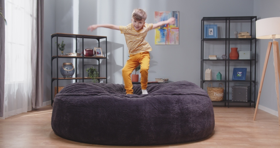 Pretty boy in yellow clothes jumping on fluffy sofa. Little happy boy in isolation. Coronavirus home quarantine. Slowmotion video.