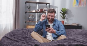 Close up portrait of adult man on sofa with smartphone in his hands listenng to music in headphones. Man dancing on couch. Male scrolling social media on gadget. Happiness concept. Slow motion video.