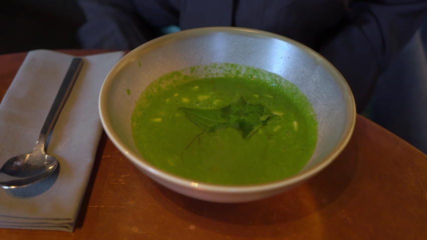 A young woman stirs soup with a spoon. A girl in a restaurant or cafe eats lunch or dinner. Vegetable Italian minestrone soup, green. Tasty meal in Italy. Female takes spoon from napkin.