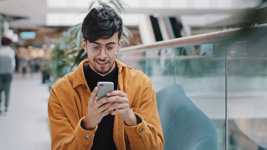 Young indian guy sitting looking at smartphone screen reading email receiving unexpected good news rejoicing smiling admiring successful online shopping at good discount wins lottery winning prize Royalty-Free Stock Footage #1087217777