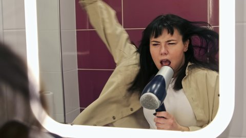 Funny woman in the bathroom dancing and emotionally singing using the hair dryer as a microphone in front of the mirror. Getting ready at home, music concept.