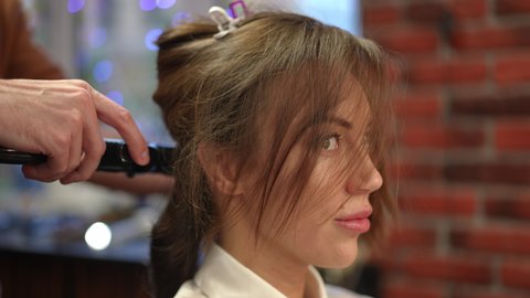 Side view close-up happy young beautiful Caucasian bride in beauty salon curling hair on wedding morning. Portrait of gorgeous woman doing hairstyle in hairdressing salon indoors. Slow motion