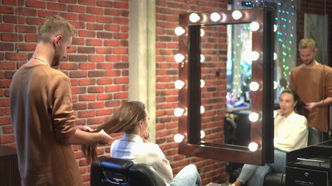Side view young male hairstylist examining long hair of female client sitting in armchair in beauty salon. Caucasian man thinking planning hairdressing indoors reflecting in mirror. Style and beauty