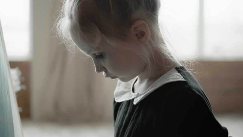 Close-up of a little seven-year-old girl in a stylish black dress, painting on a canvas with paints and brushes