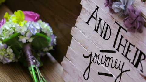 Sign with the text "And Their Journey Begins" sitting on a pew in a church next to a bouquet of flowers.