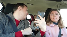 Portrait of beautiful married couple and their happy children waving hello to their family on mobile phone screen while sitting in car during family trip.