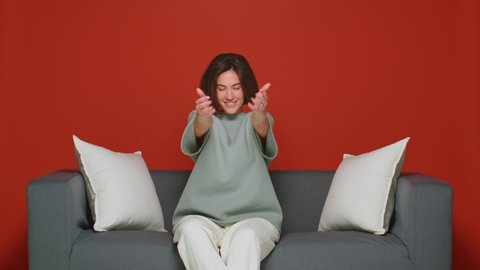 A young pretty girl is sitting on the couch at home and beckoning to her with a beckoning gesture. Isolated on a red background.
