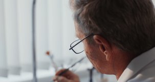 An orthodontist - a mature man in glasses manually applies a coating on a denture using a thin brush. Close-up, view from behind. Video in 4k, red komodo