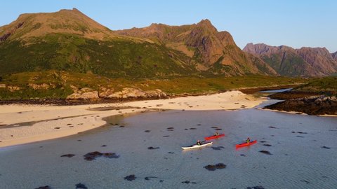 Aerial view around kayakers paddling in shallow waters with beach and mountain background, sunset in Norway - circling, drone shot