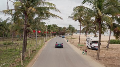 Sanya , Hainan island , China - 02 09 2022: Tesla Model 3 driving on tropical road by the beach with numerous camper vans parked on the roadside. Hainan island, China.