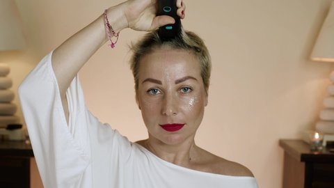 A woman with red lipstick shaves her head with a clipper at home