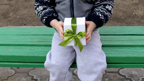 little kid is sitting on a green park bench with a gift box in hand or on the seat. white box with green ribbon. present to his beloved girl. waiting the girl friend. boy is alone outside. march 8, wo