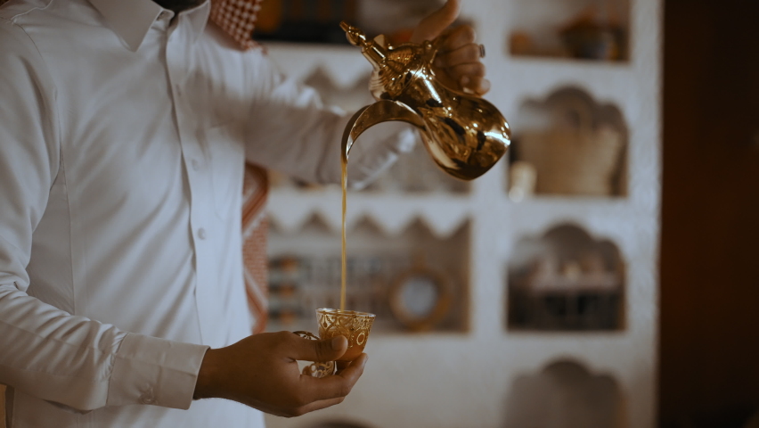 Saudi man wearing Thoub pouring coffee from dallah into finjal with bokeh background indoor and old home fire place in the background  | Shutterstock HD Video #1087253867
