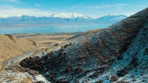 A dirt road through a canyon leading to a town in the valley with snow-capped mountains across a lake - aerial pullback view