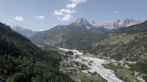 Frontal drone video advancing between the mountains over the Rragam i Shalese village in the Valbona valley and the Rrogam i Shales river. Clear sky with few clouds and few buildings.
