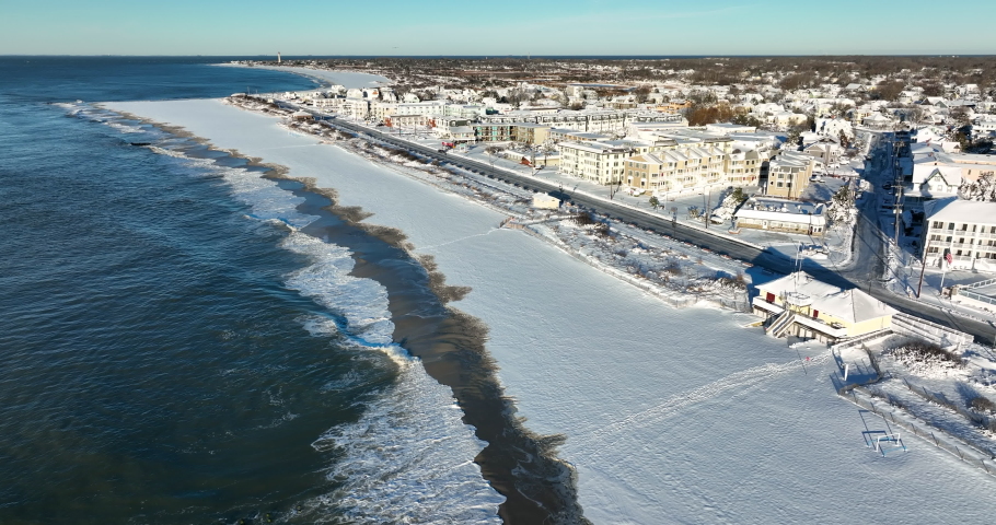 Atlantic Ocean beach covered in winter snow. Tourist town in off-season. Beachfront hotels with water view. Waves crash on sandy beach.