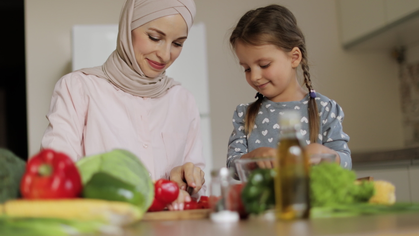 Pretty muslim mother and daughter preparing tasty food at kitchen. Mommy teaching lovely kid to cook. Happy mom and loving child spending time together at home. Healthy meal and dinner preparation