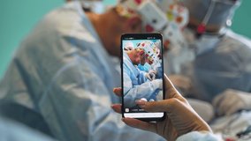 Female hand holding a telephone and taking video of operating surgeons. Close up. Backdrop in blur.
