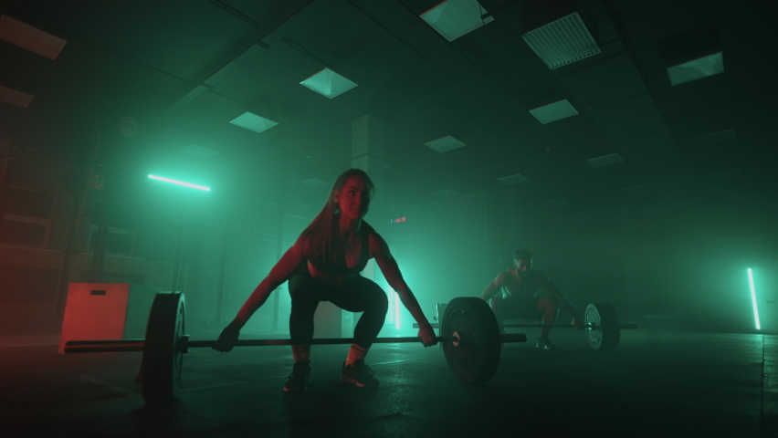 A man and a woman in a colored neon red and blue light in slow motion lift heavy barbells over their heads. Strength and power training with heavy weight. The concept of athletic and strong couples Royalty-Free Stock Footage #1087267907