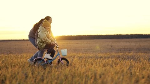 childhood dream. woman mother teaches child to ride bike in the park. Driving lesson for kid from mom at sunset. parent helps child to keep balance and pedal. Kid learns steer bike sun. happy family